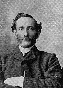 Portrait of Archibald Meston, a man with a moustache wearing a suit and crossing his arms.